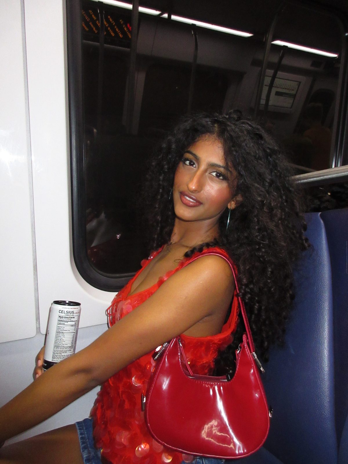Jerusha on a train in a red sequin top, holding a drink with a red shoulder bag
